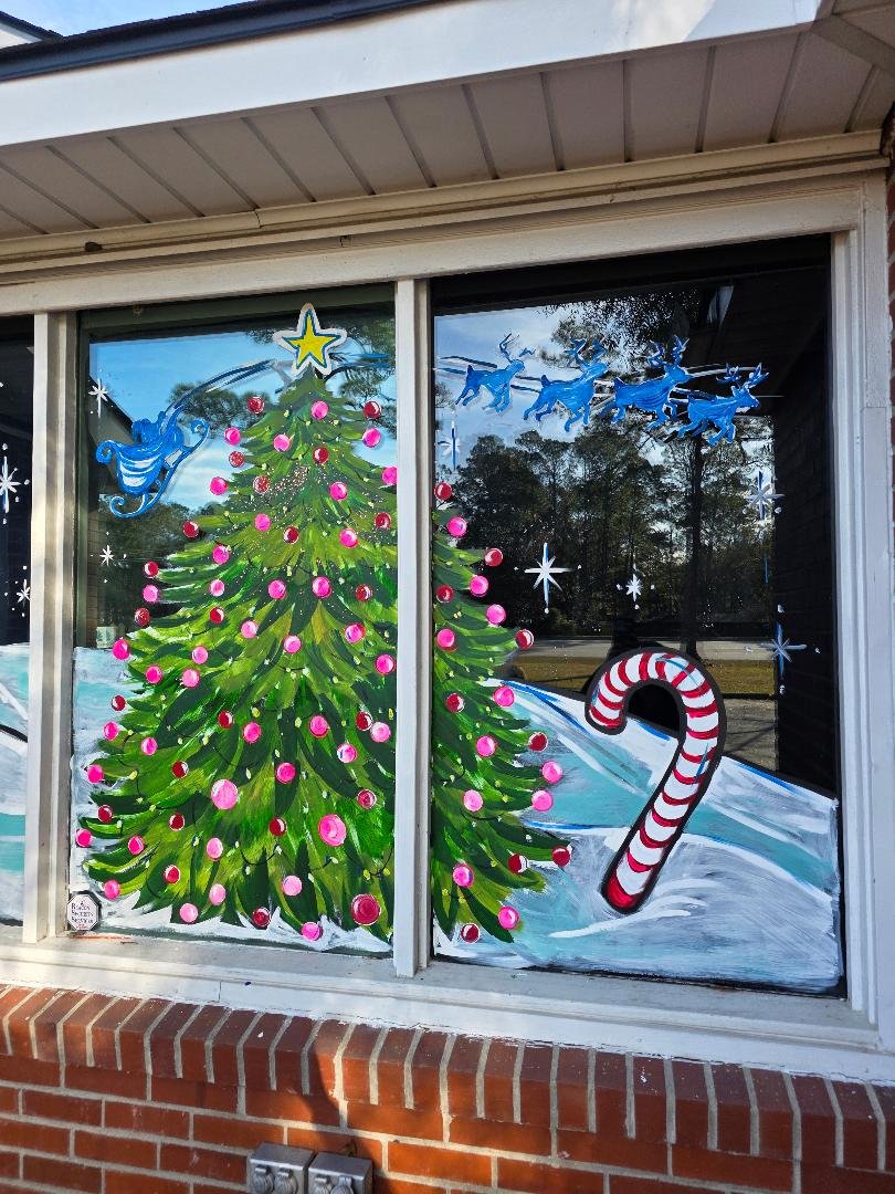 Decorated window with a painting of a Christmas tree with pink ornaments, a golden star on top, behind snow-covered ground, a candy cane in the snow, blue reindeer, and white snowflakes.