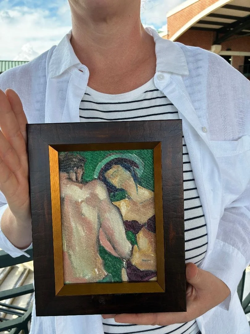 Person wearing a white shirt and striped top holding a framed painting of two nude women with dark hair, standing close together, against a green background.