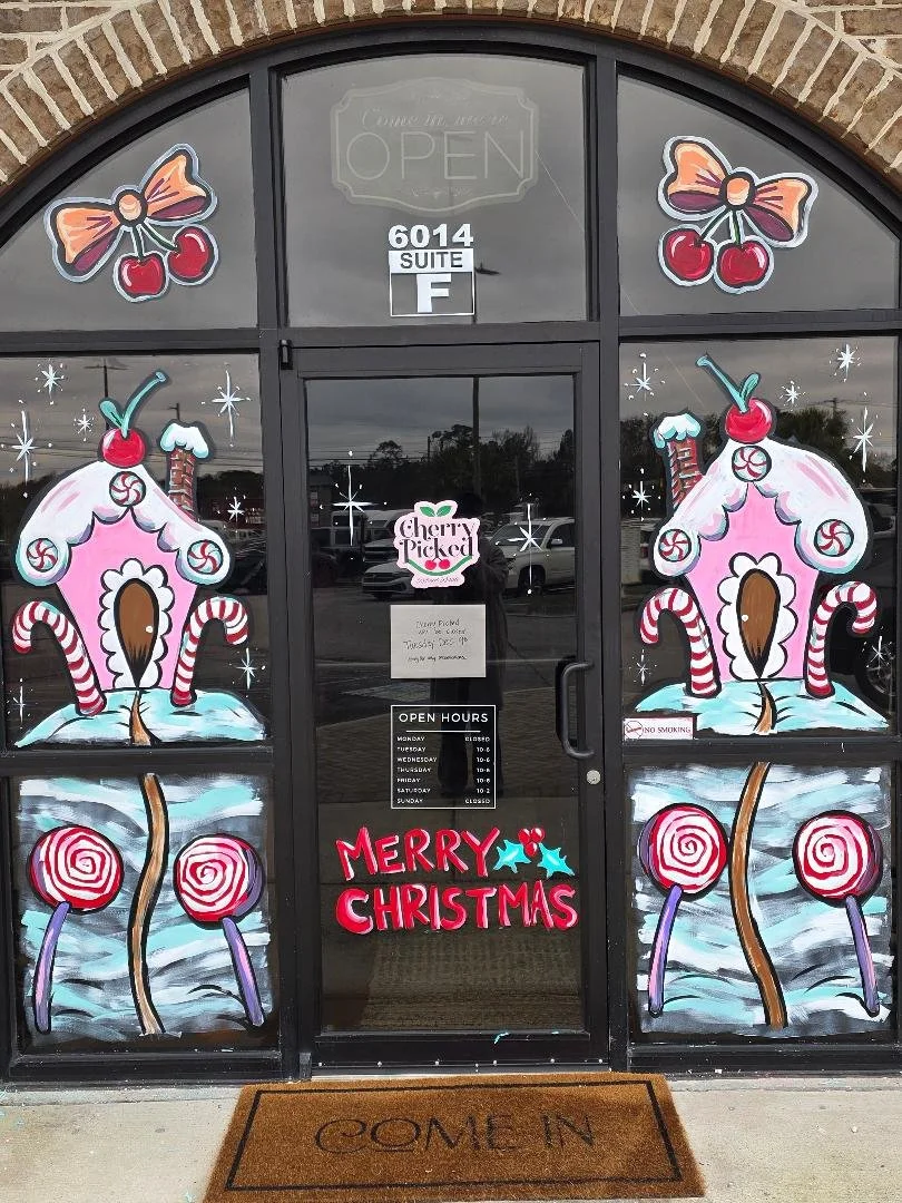 Storefront window painting Christmas-themed  including candies, lollipops, gingerbread houses, and bows, with a "Merry Christmas" sign and an open sign on the door.
