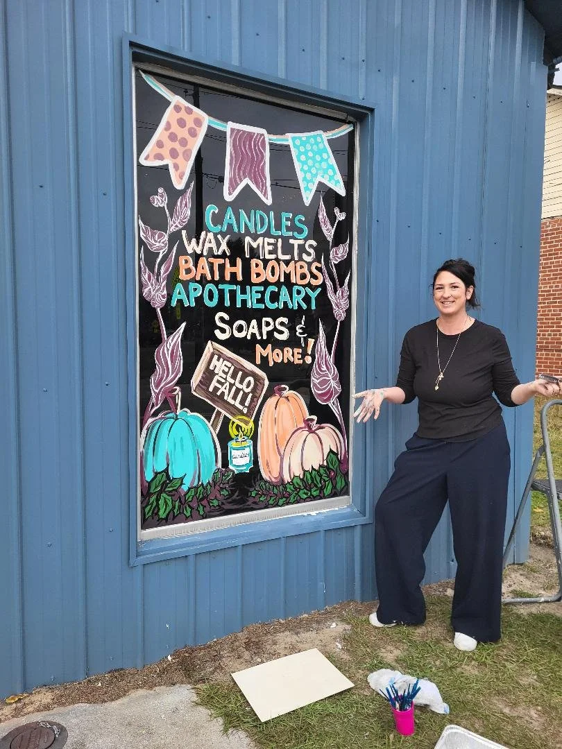 colorful fall-themed window painting advertising candles, wax melts, bath bombs, apothecary, soaps, and more, with pumpkins and decorative leaves.