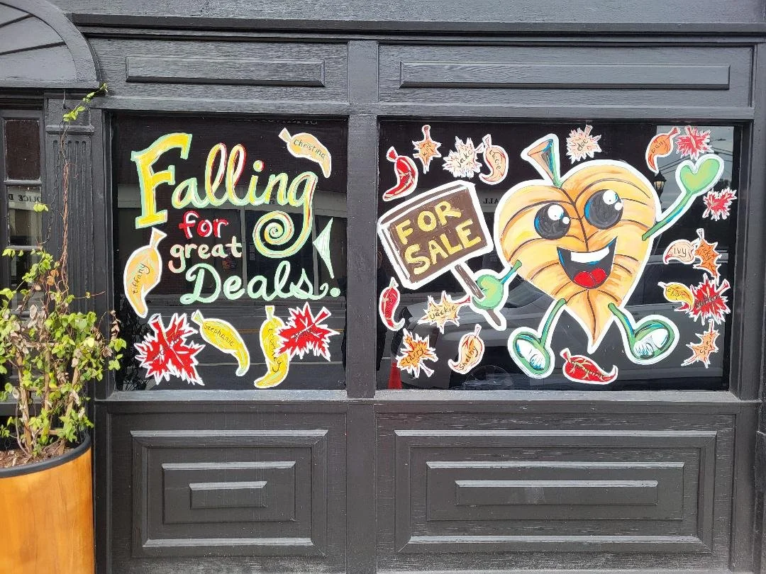 Decorative window display with signs and cartoon pumpkin character celebrating fall. Text reads "Falling for great Deals!" and "FOR SALE" with colorful autumn leaves and chili peppers.