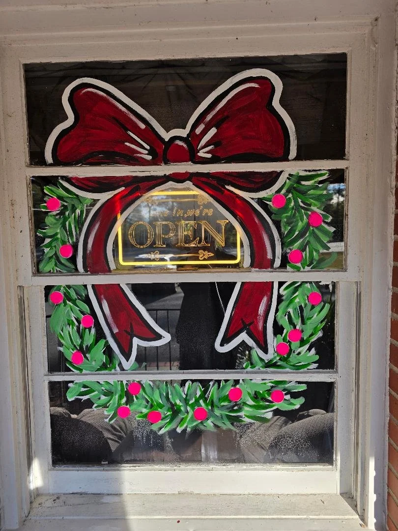 Decorative Christmas wreath with a large red bow and an illuminated sign saying "We are OPEN" on a window.