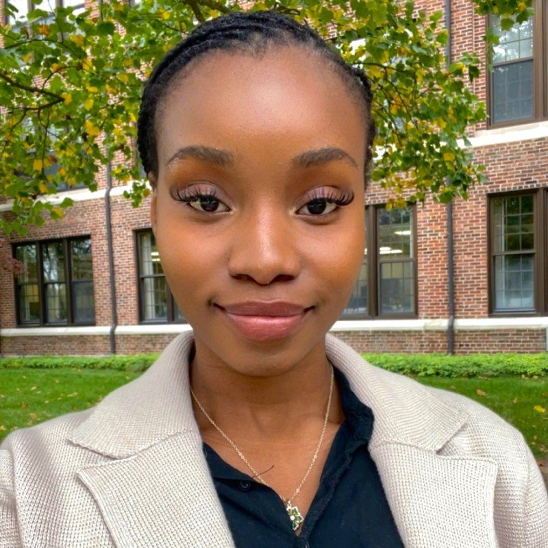 A young woman with braided hair, wearing a beige blazer over a black shirt, standing outdoors with a brick building and green trees in the background.