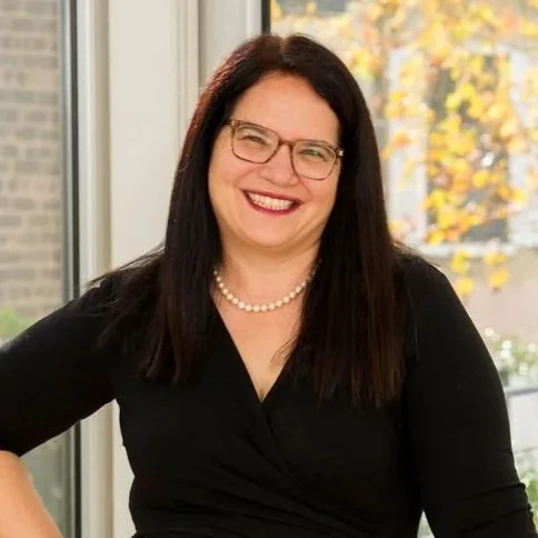 A woman with long dark hair, glasses, red lipstick, and a red bead necklace with a white circular pendant, smiling at the camera.
