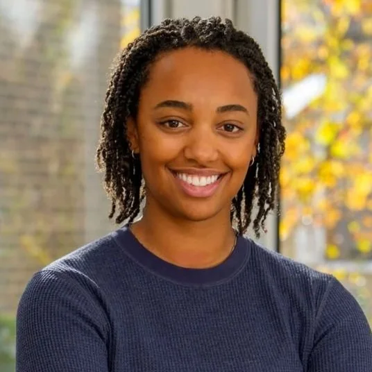A young woman with curly dark hair and a light brown complexion, wearing a purple top and smiling softly at the camera.