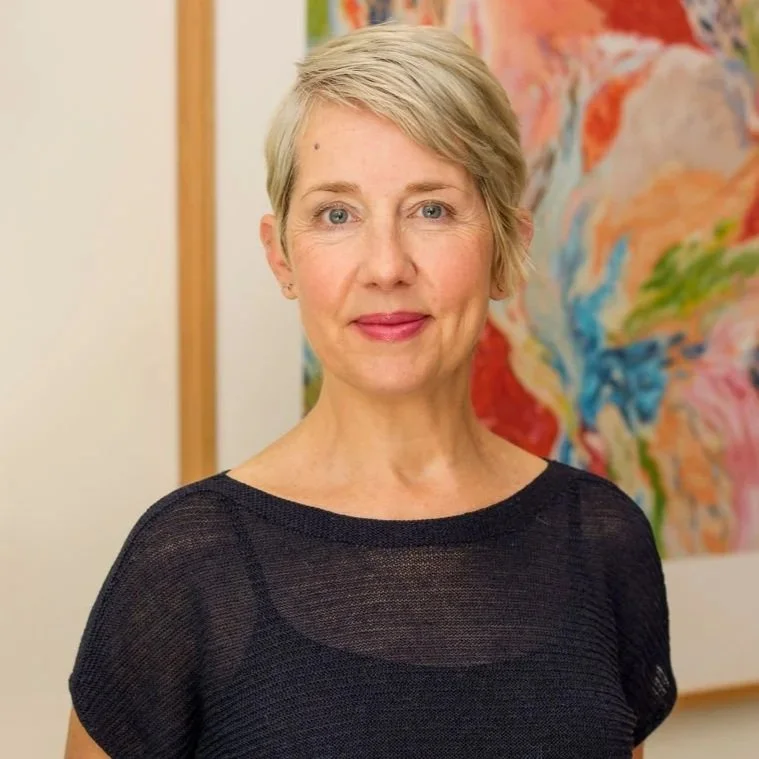 A smiling woman with short, curly blonde hair, wearing a striped shirt, standing against a plain white background.