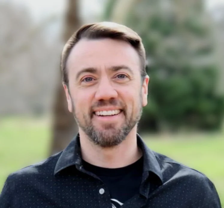 an image of Elijah smiling at the camera, wearing a button-down shirt over a t-shirt.