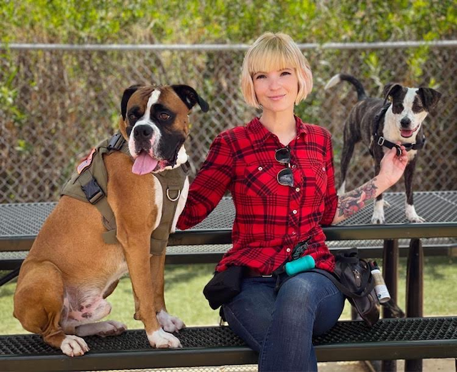 A young woman with blonde hair and a red plaid shirt sitting on a park bench outdoors, with two dogs, one on her left and one on her right. The woman is smiling, and the dogs appear happy, with one sitting and the other standing.