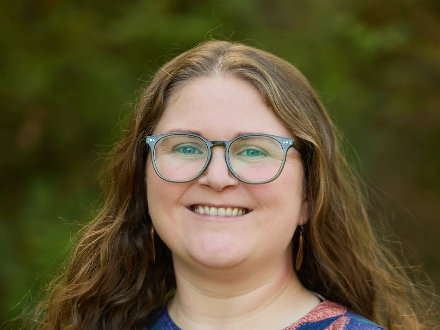 A smiling woman with long brown hair, wearing clear glasses and a colorful striped top, outdoors with green foliage in the background.