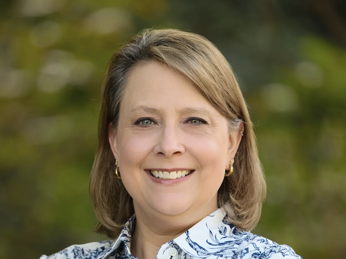 Smiling middle-aged woman with blonde hair wearing a blue-and-white floral blouse outdoors