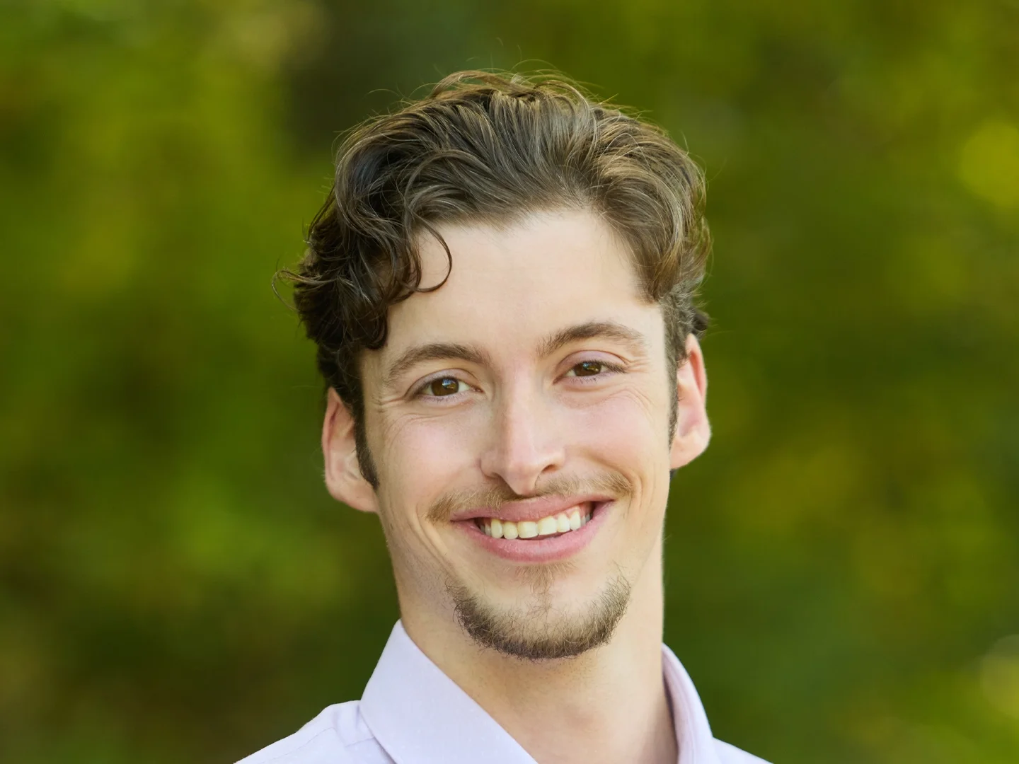 Smiling young man with curly brown hair and goatee outdoors against a blurred green background
