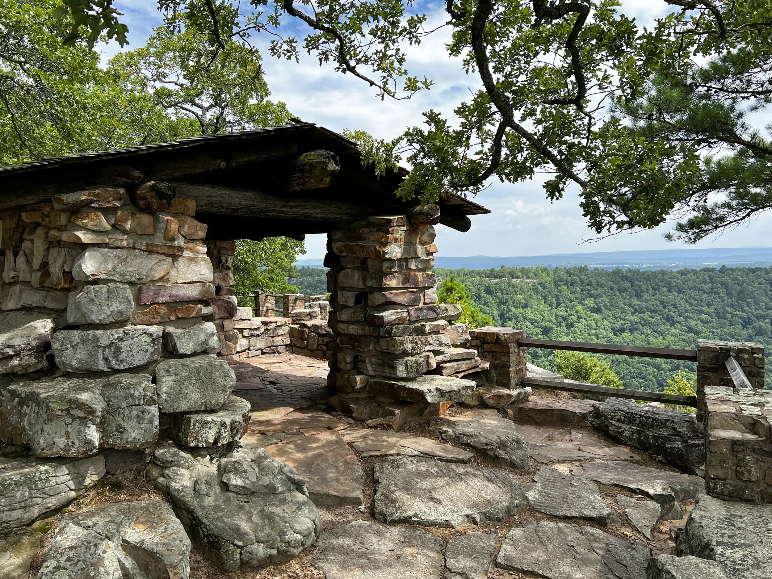 Stone mountain shelter with a log roof overlooking a forested valley and distant mountains