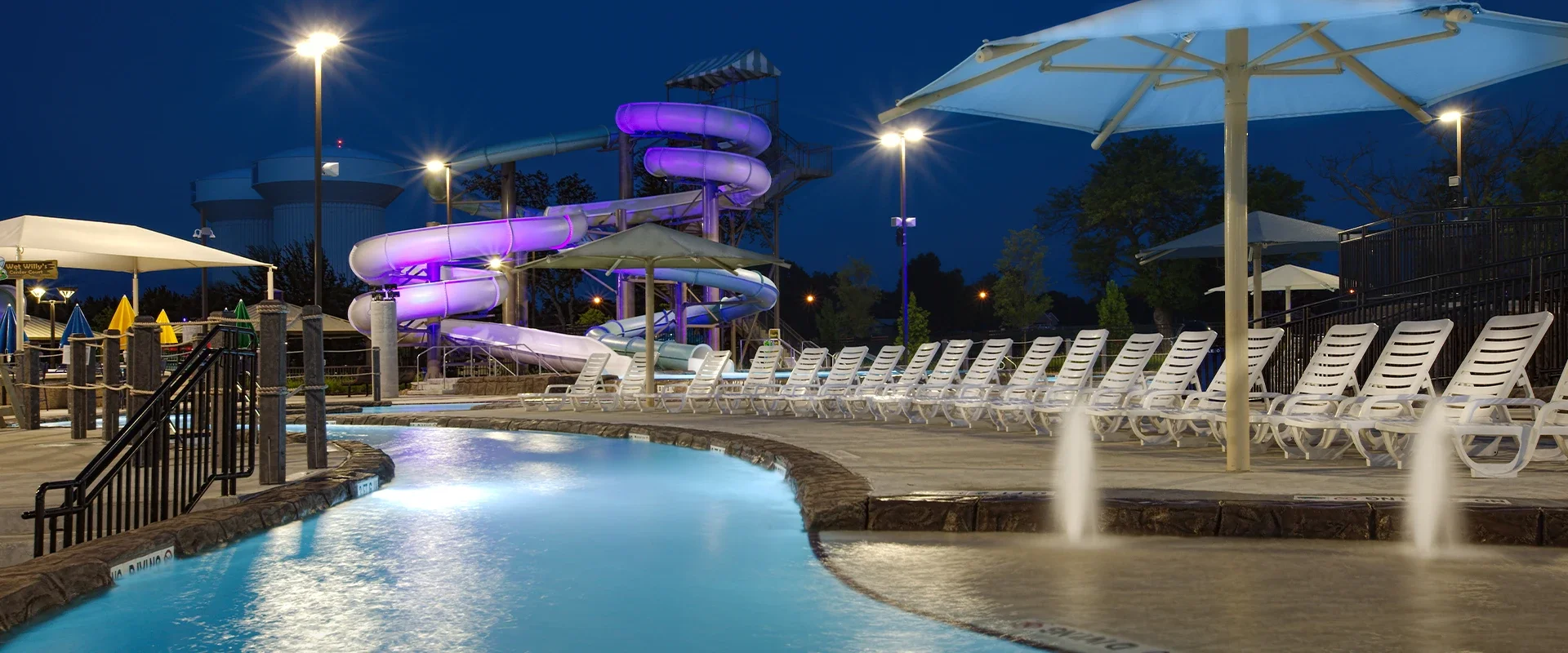 Nighttime view of a water park with purple-lit waterslides, pool, fountains, umbrellas, and rows of lounge chairs.
