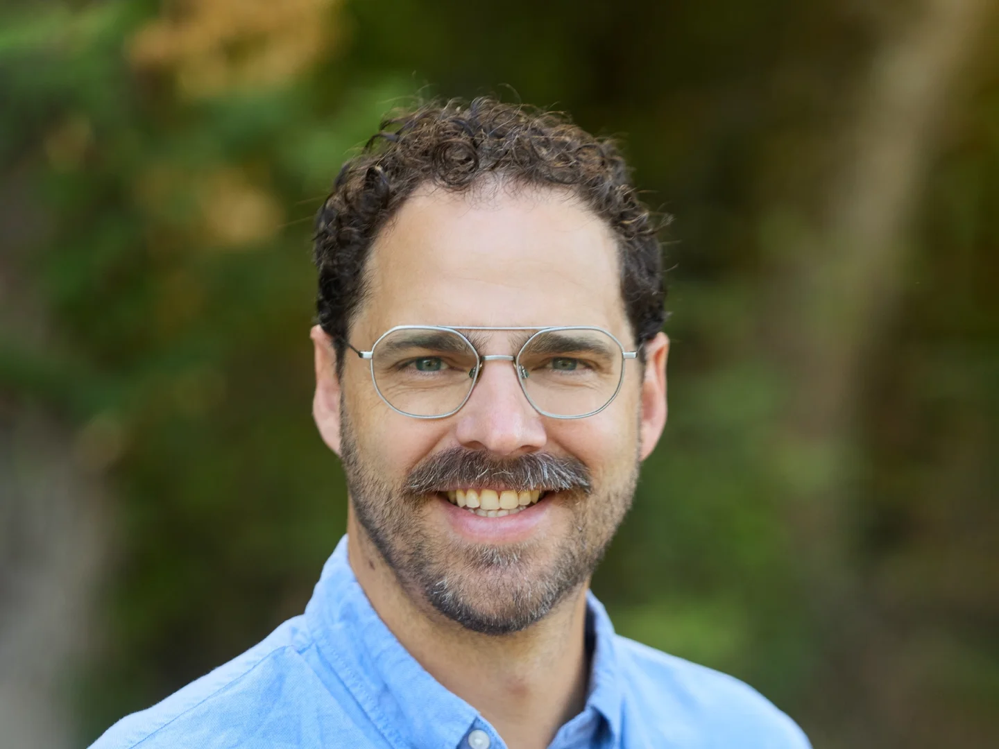 A man with glasses and a beard smiling outdoors with trees in the background.