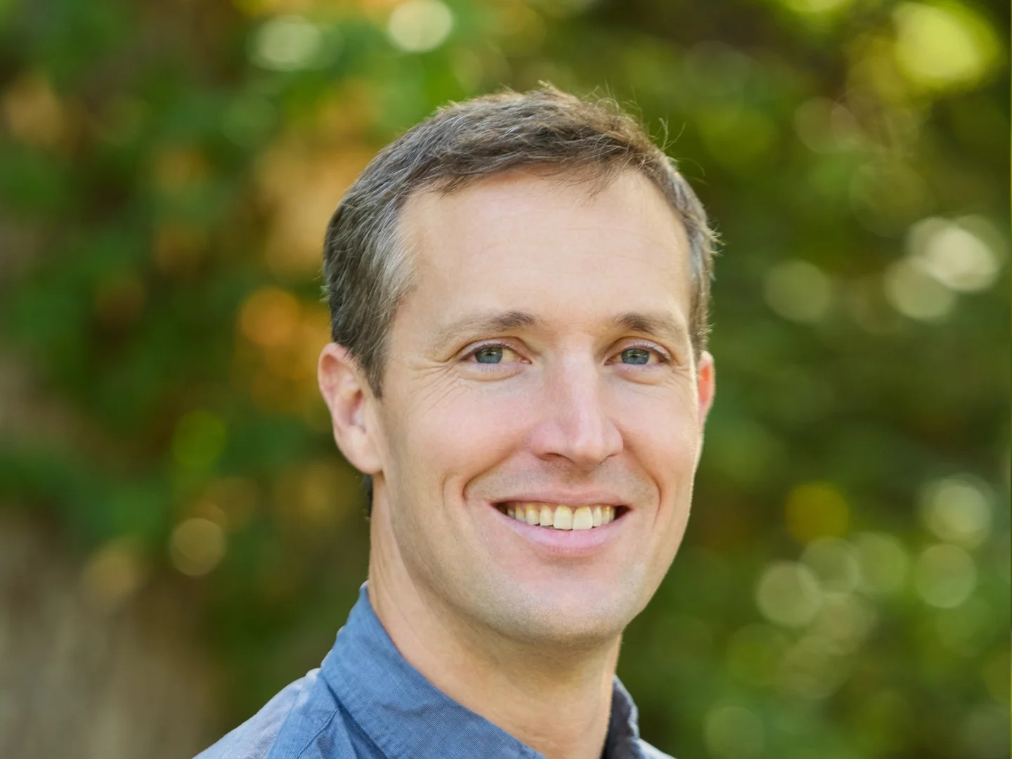 A smiling man with light skin, brown hair, and blue eyes, wearing a checkered shirt, outdoors with green foliage in the background.
