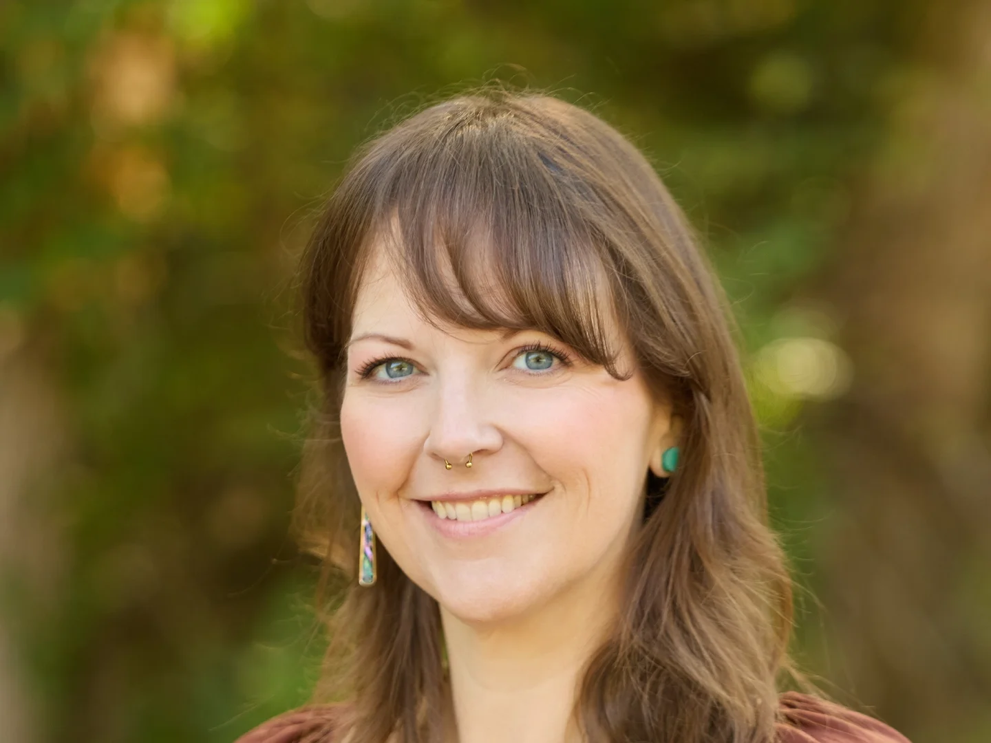 Close-up of a woman with long brown hair, blue eyes, and a bright smile, outdoors with green foliage in the background.