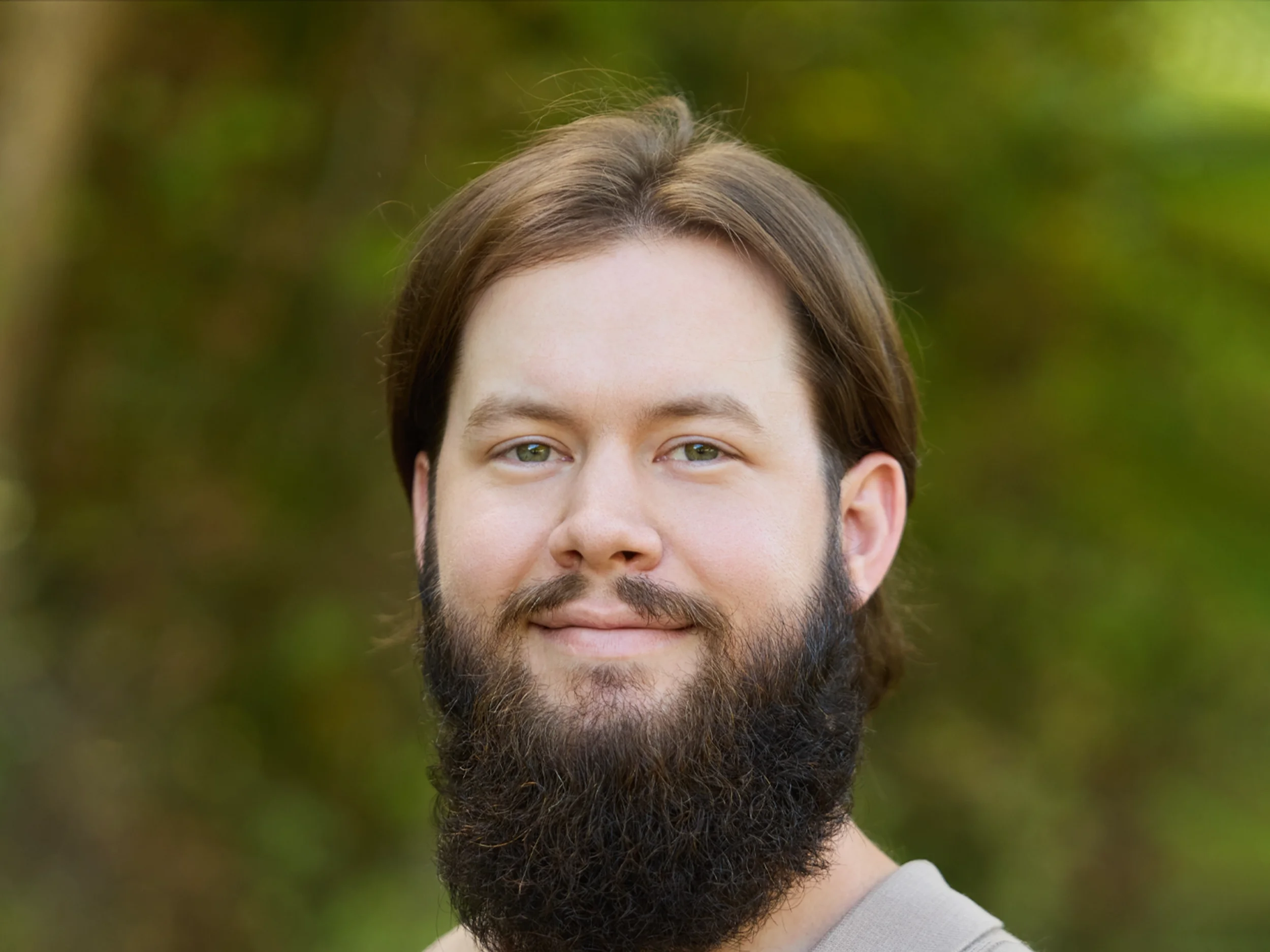 A man with a beard and blue eyes smiling outdoors with green foliage in the background.