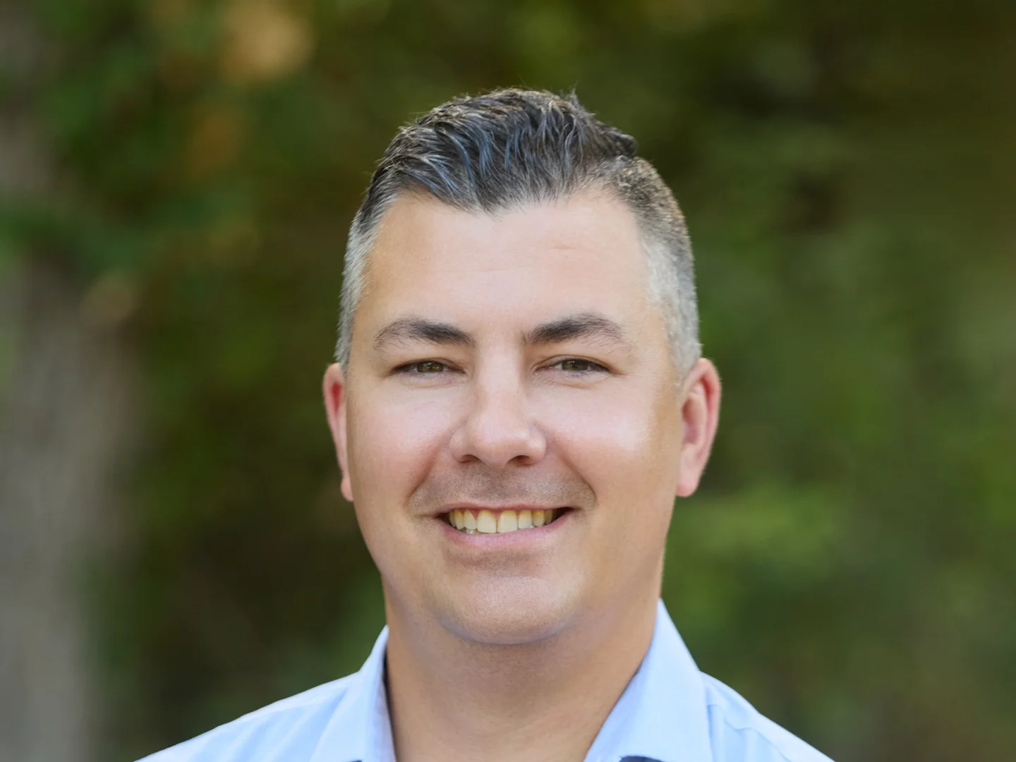 A smiling man outdoors with a blurred green leafy background.