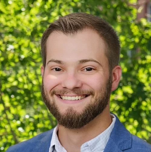 Portrait of a young man with short hair, a beard, smiling, wearing a blue blazer and white shirt, outdoors with green foliage in the background.