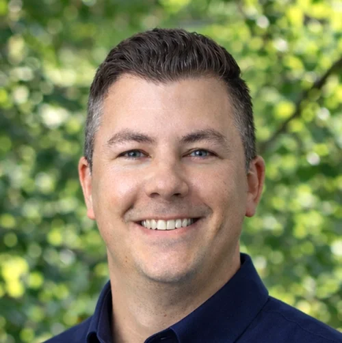 A smiling man with short dark hair and blue eyes, standing outdoors with a background of green leaves and trees.