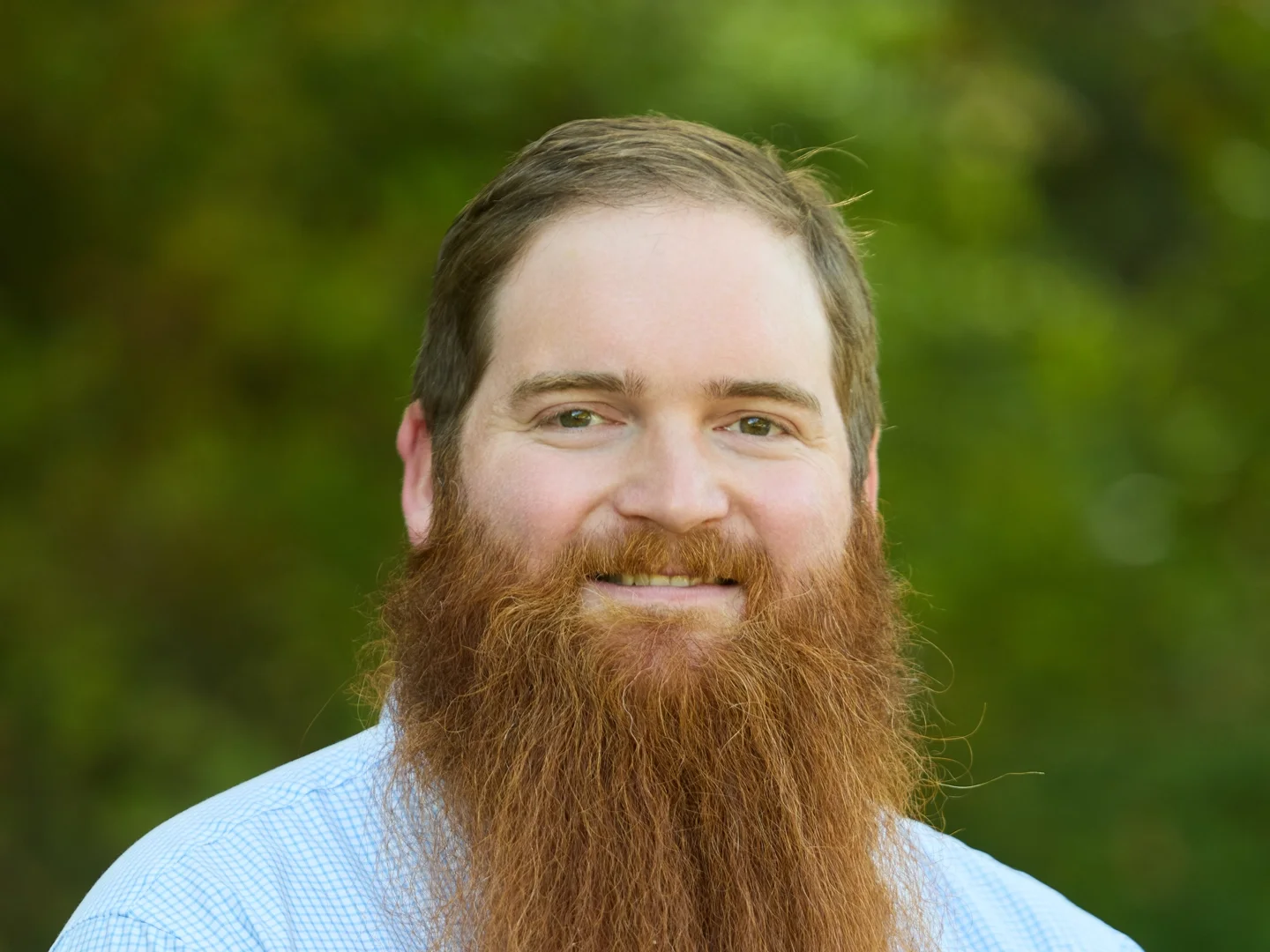 Smiling bearded man in a light blue shirt posing outdoors against a blurred green background