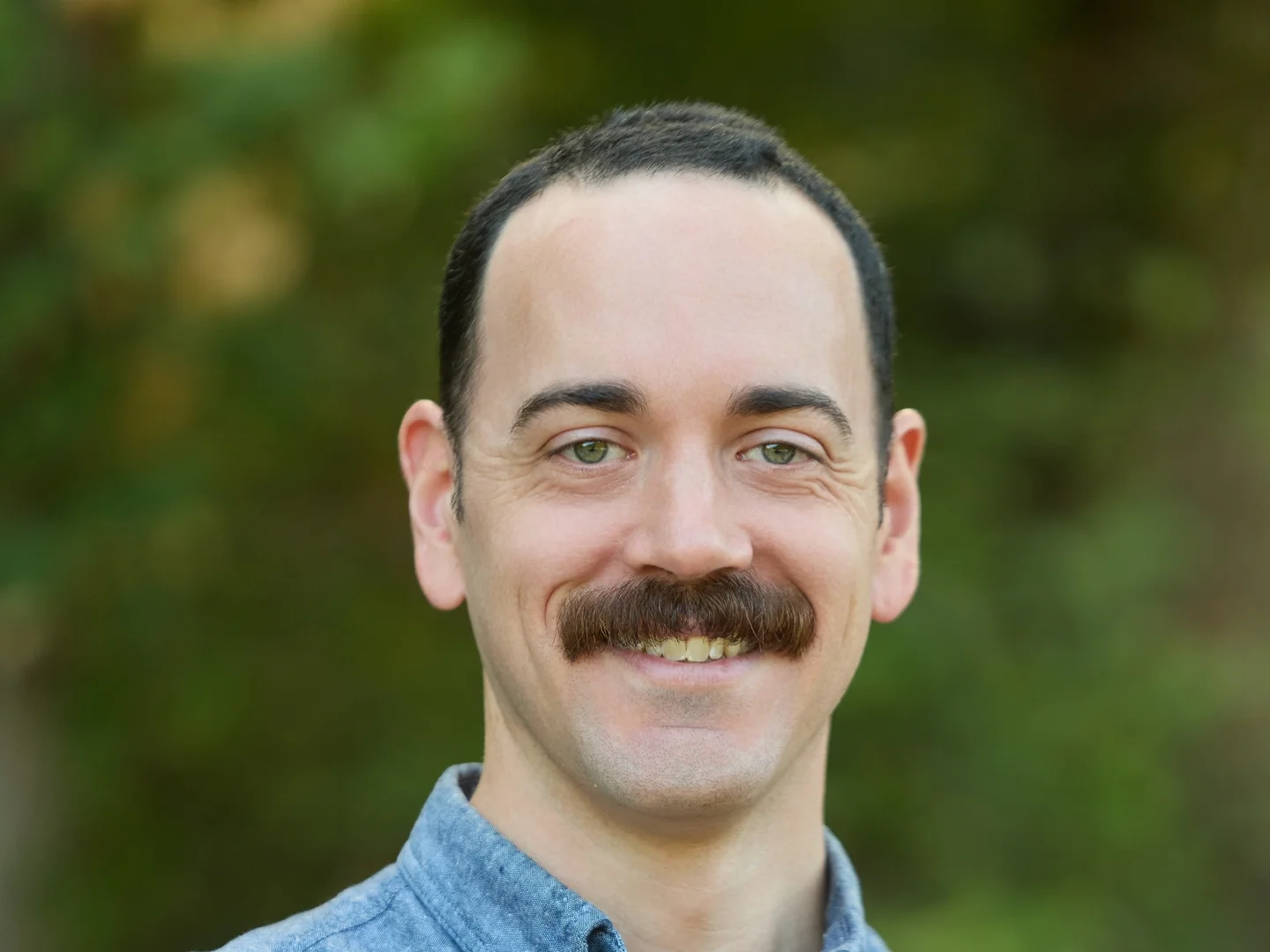 A man with dark hair, a mustache, and a big smile outdoors with green leafy trees in the background.