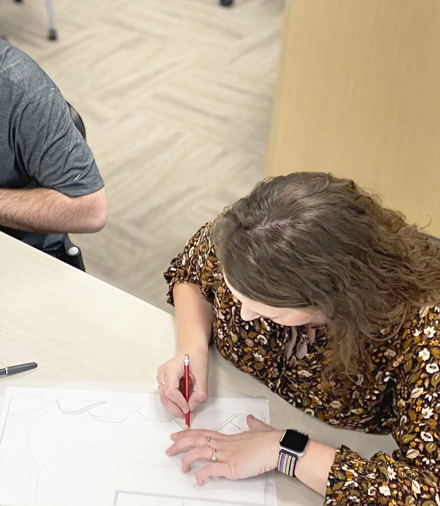 Woman in a floral blouse sketching on paper with a red pencil at a table, viewed from above