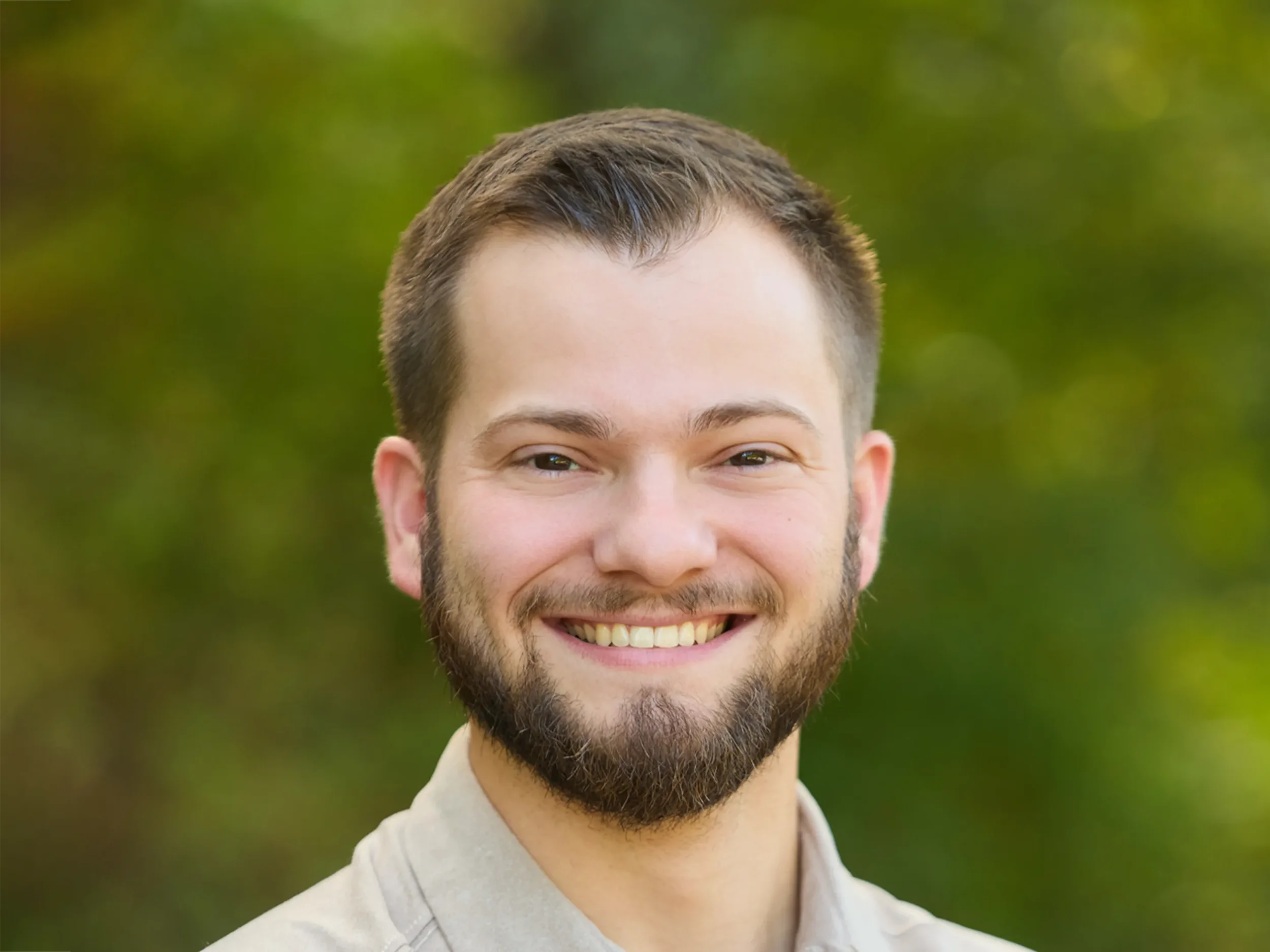 Headshot of a man with short brown hair and beard, smiling, in front of a green leafy background.