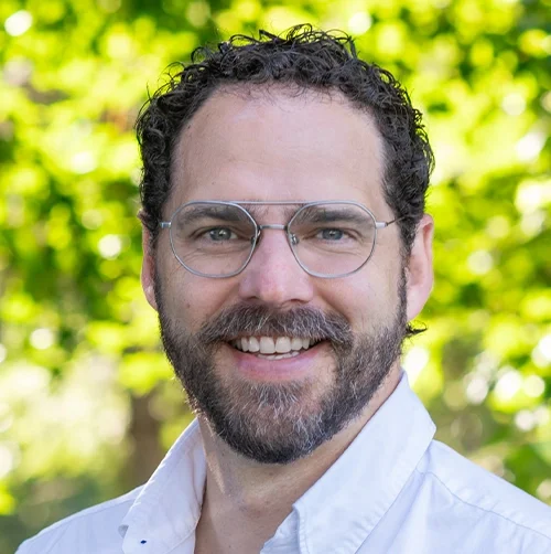 A man with glasses and a beard smiling outdoors with trees in the background.