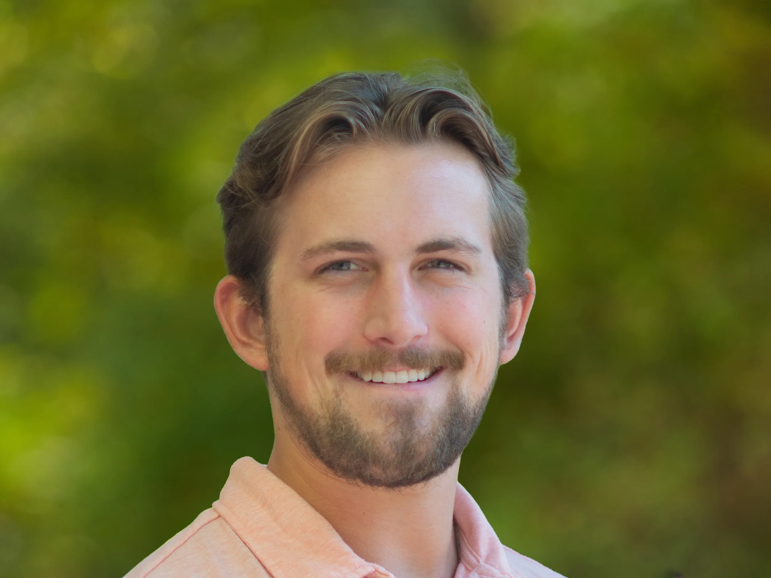 Smile of a young man with brown hair and a beard outdoors with greenery in the background.