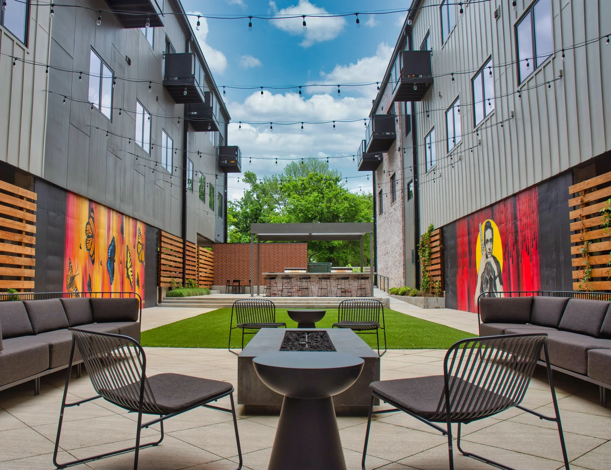 Modern courtyard patio with lounge seating, fire pit, string lights, murals, and outdoor bar between buildings