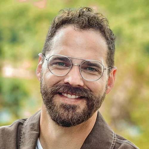 A smiling man with glasses and a beard outdoors with a blurred green background.