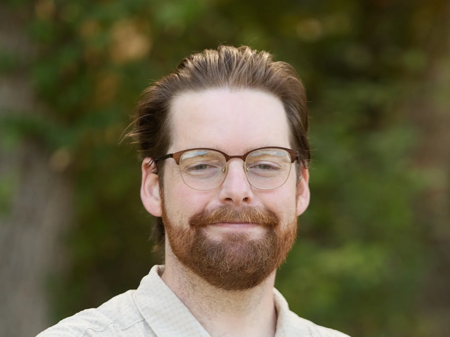 Portrait of a smiling bearded man wearing glasses outdoors with a blurred green forest background