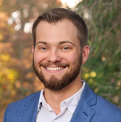 Portrait of a young man with short hair, a beard, smiling, wearing a blue blazer and white shirt, outdoors with green foliage in the background.