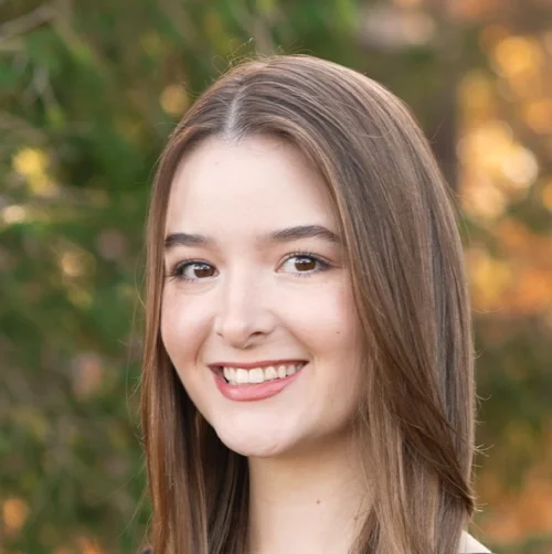 Young woman with long brown hair smiling outdoors with trees and warm sunlight in the background.