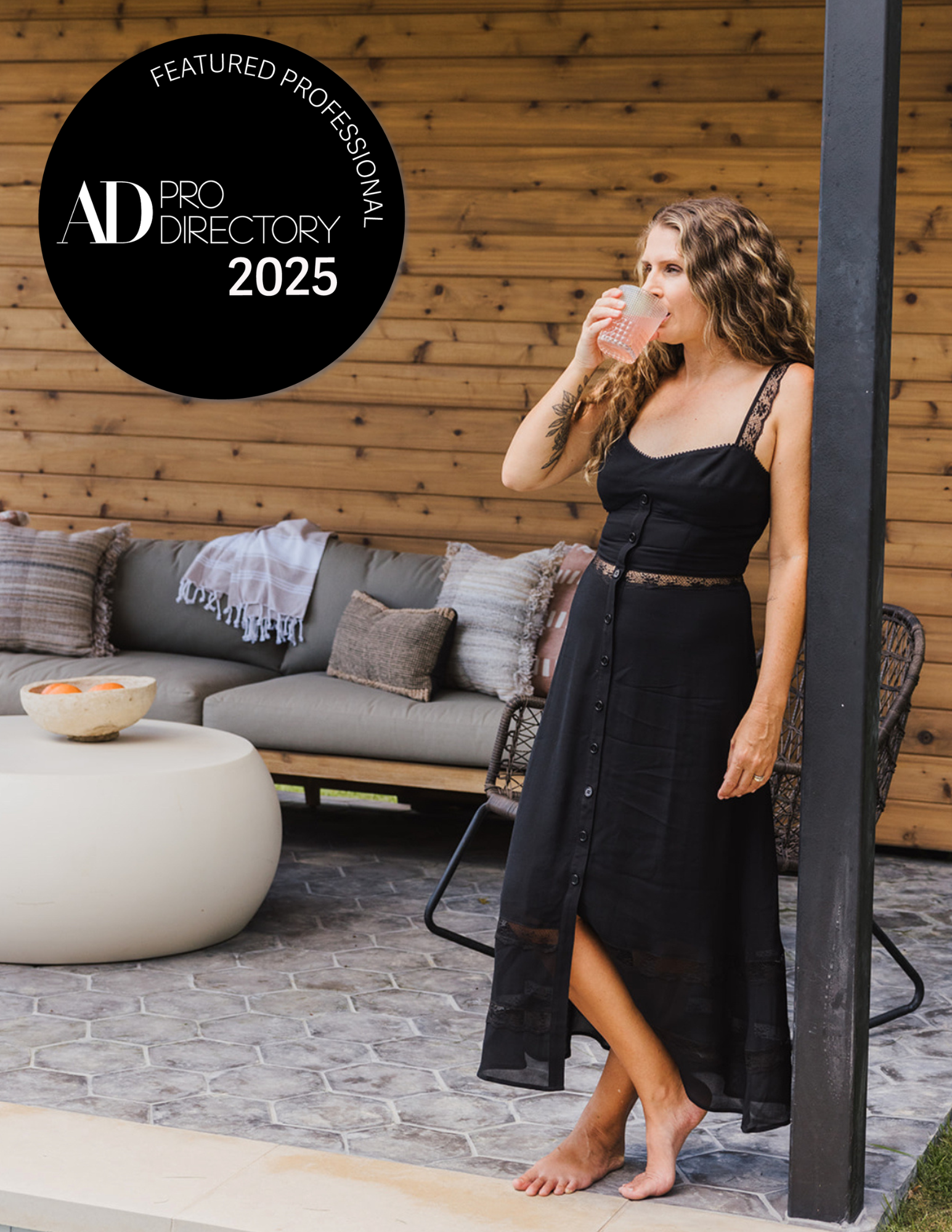 Woman in a black dress drinking a pink beverage outside on a patio with a wood-paneled wall and a sofa with pillows in the background, featuring a badge indicating recognition in the 2025 AD Professional Directory.