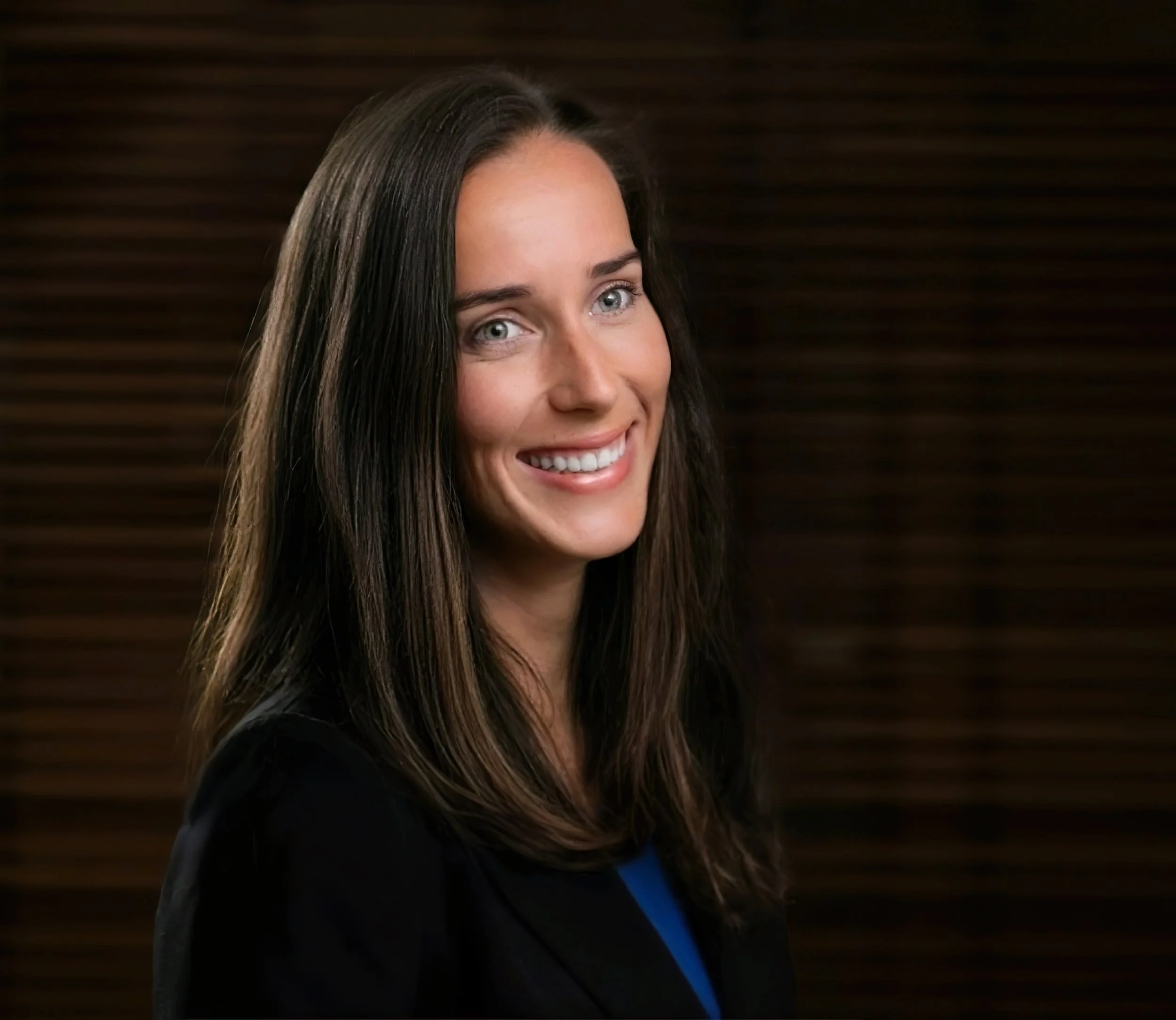 Smiling woman with long brown hair, wearing a black blazer and blue top, photographed against a softly lit dark background.