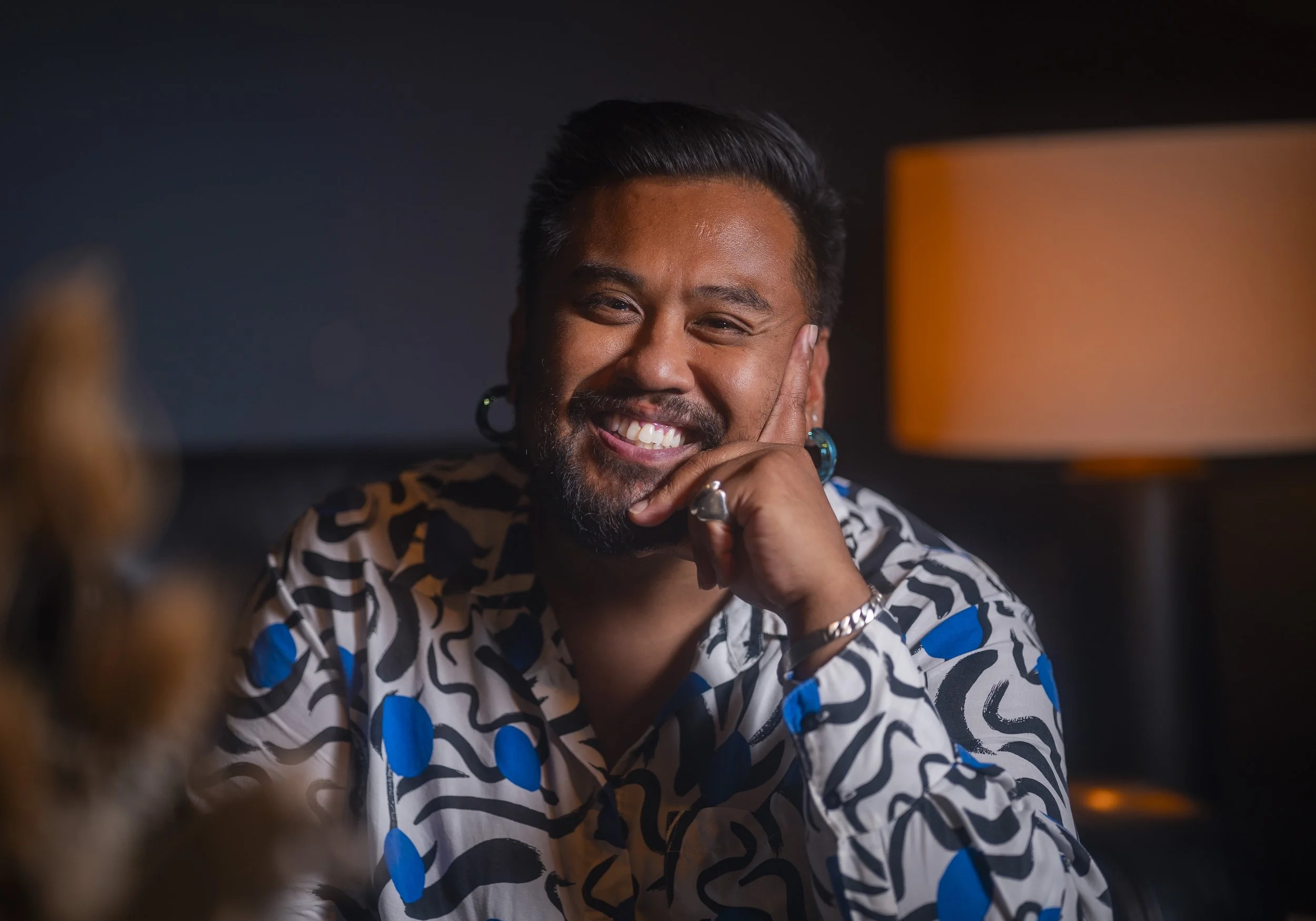 Smiling person with dark hair and beard, wearing a patterned shirt with blue accents, resting their chin on their hand, with warm indoor lighting in the background.
