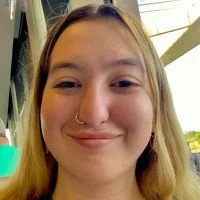 Smiling young woman with long blonde hair, wearing a nose ring and earrings, photographed indoors with natural light from large windows.