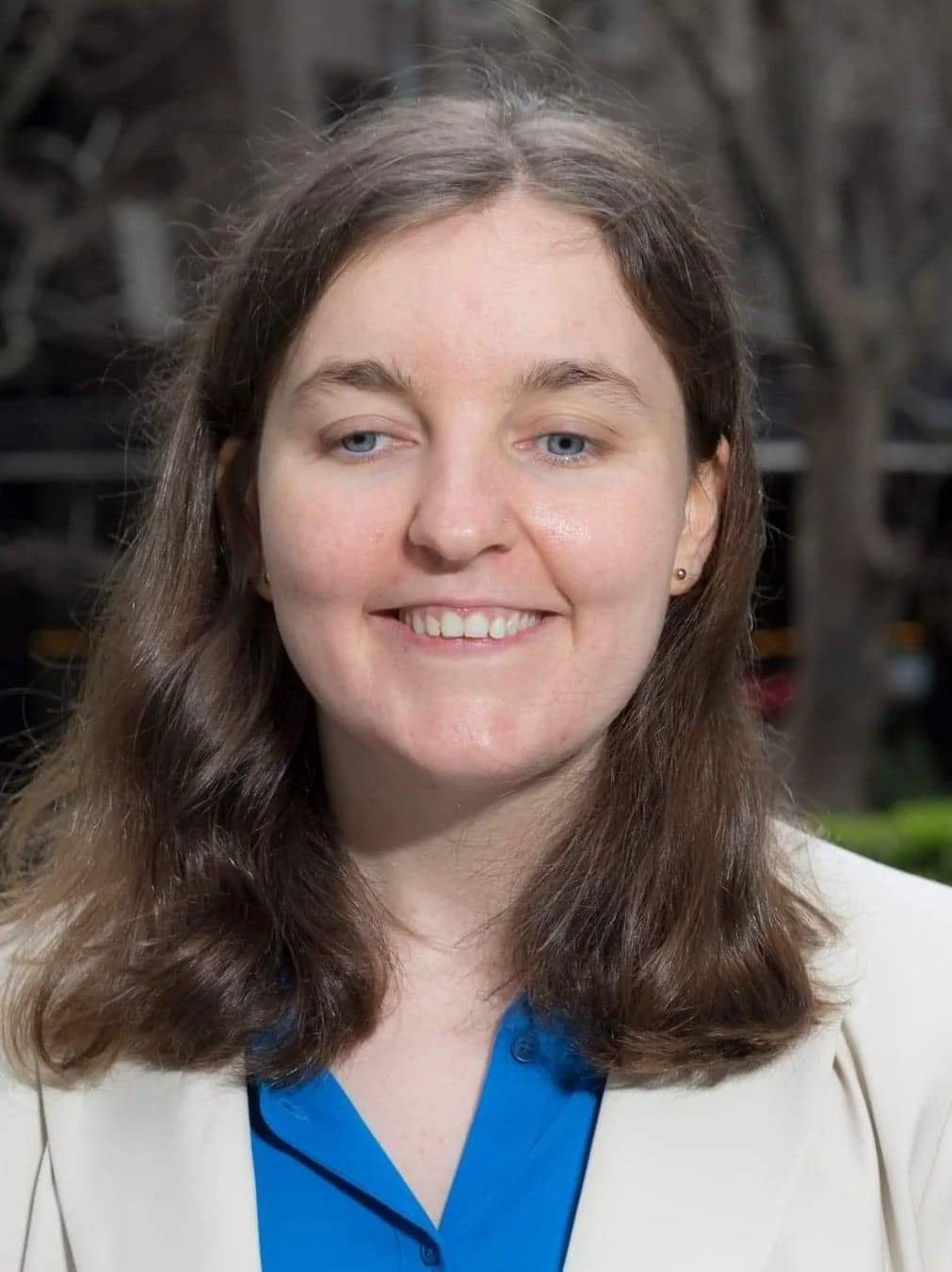 Smiling woman with shoulder-length brown hair wearing a cream blazer and blue shirt, photographed outdoors with blurred trees in the background.