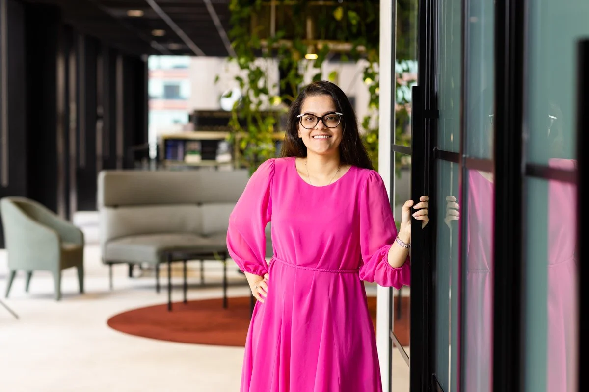 Smiling woman with long dark hair, wearing glasses and a bright pink dress, standing with one hand on her hip in a modern office space with plants and lounge seating.