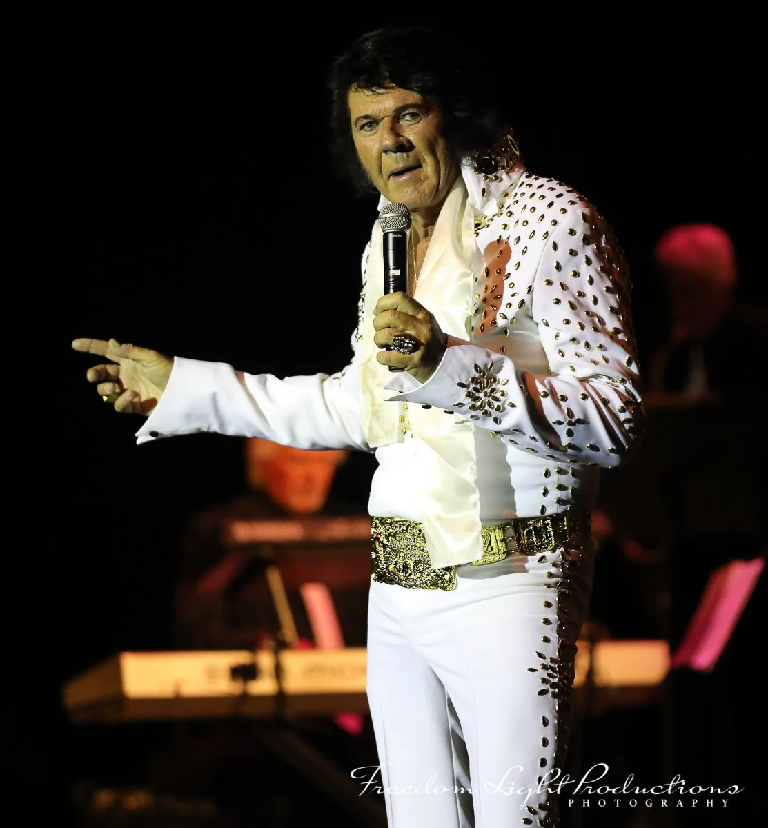 Elvis Presley performing on stage in a white sequined jumpsuit with gold accents, holding a microphone and gesturing with his left hand, with a blurred band in the background.