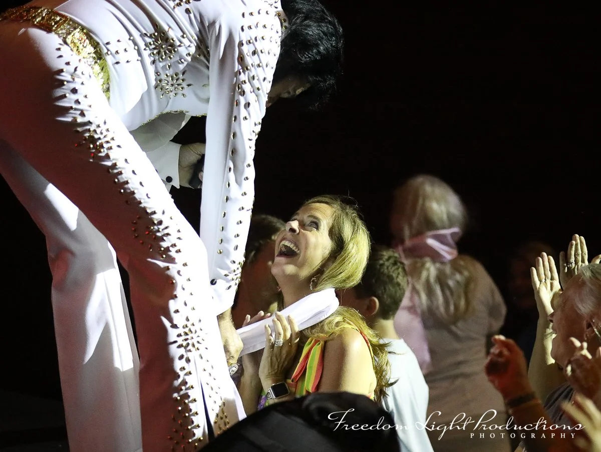 A woman with a joyful expression looking up at a man dressed as Elvis Presley, both in a crowd at a celebration.