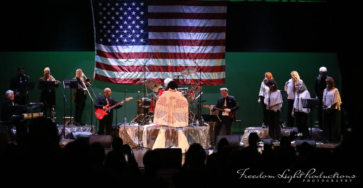 A band performing on stage with American flag backdrop, consisting of musicians playing various instruments including drums, guitars, keyboard, and singers, in a concert setting with audience silhouette in foreground.