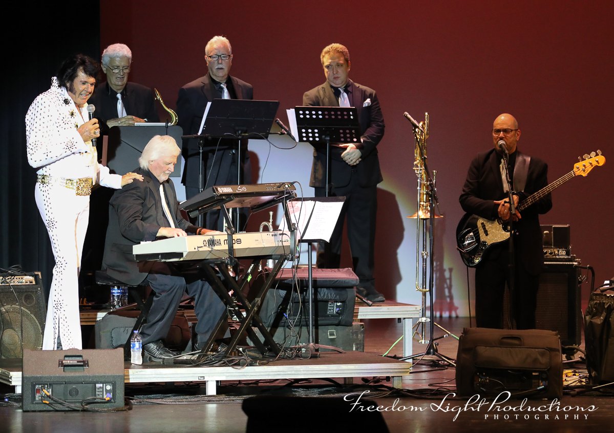 Six musicians performing on stage with musical instruments, including a keyboard, saxophone, and electric bass guitar, in a concert setting.
