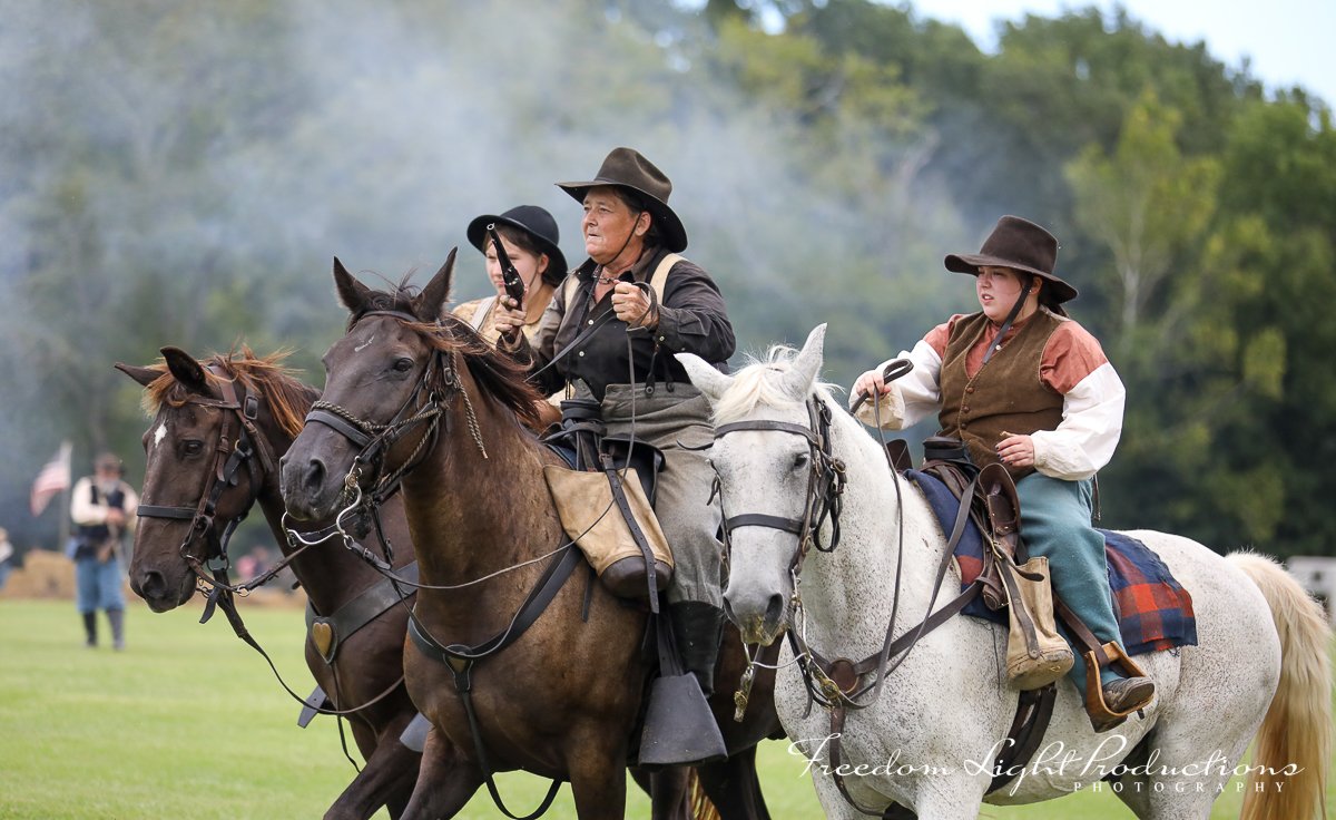 Three people in historical attire riding horses on a grassy field during a reenactment event.