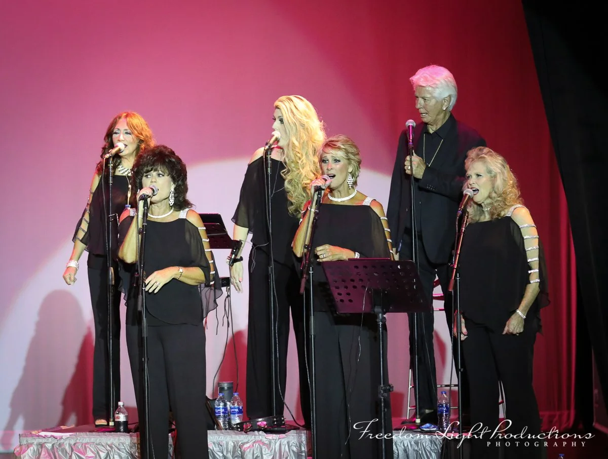 Six women and one man singing on stage with microphones, dressed in black, with a pink and red background, and stage lighting.