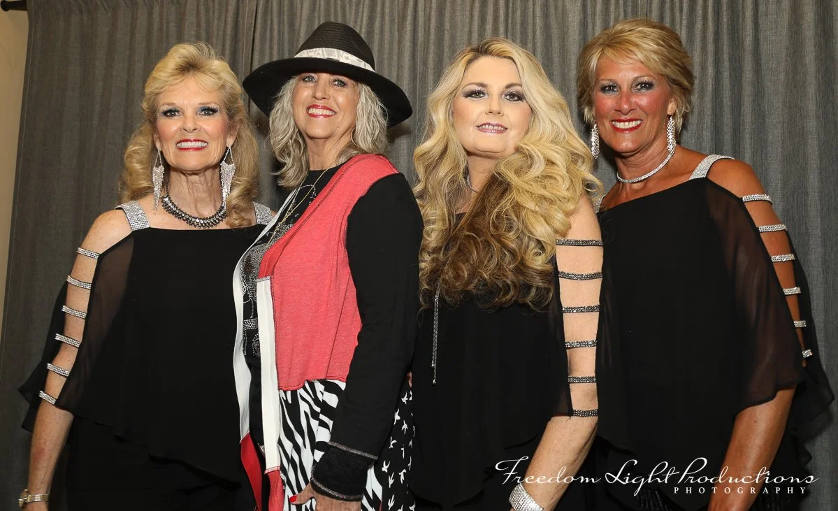 Four women dressed in black with sparkly accessories standing together in front of a curtain.