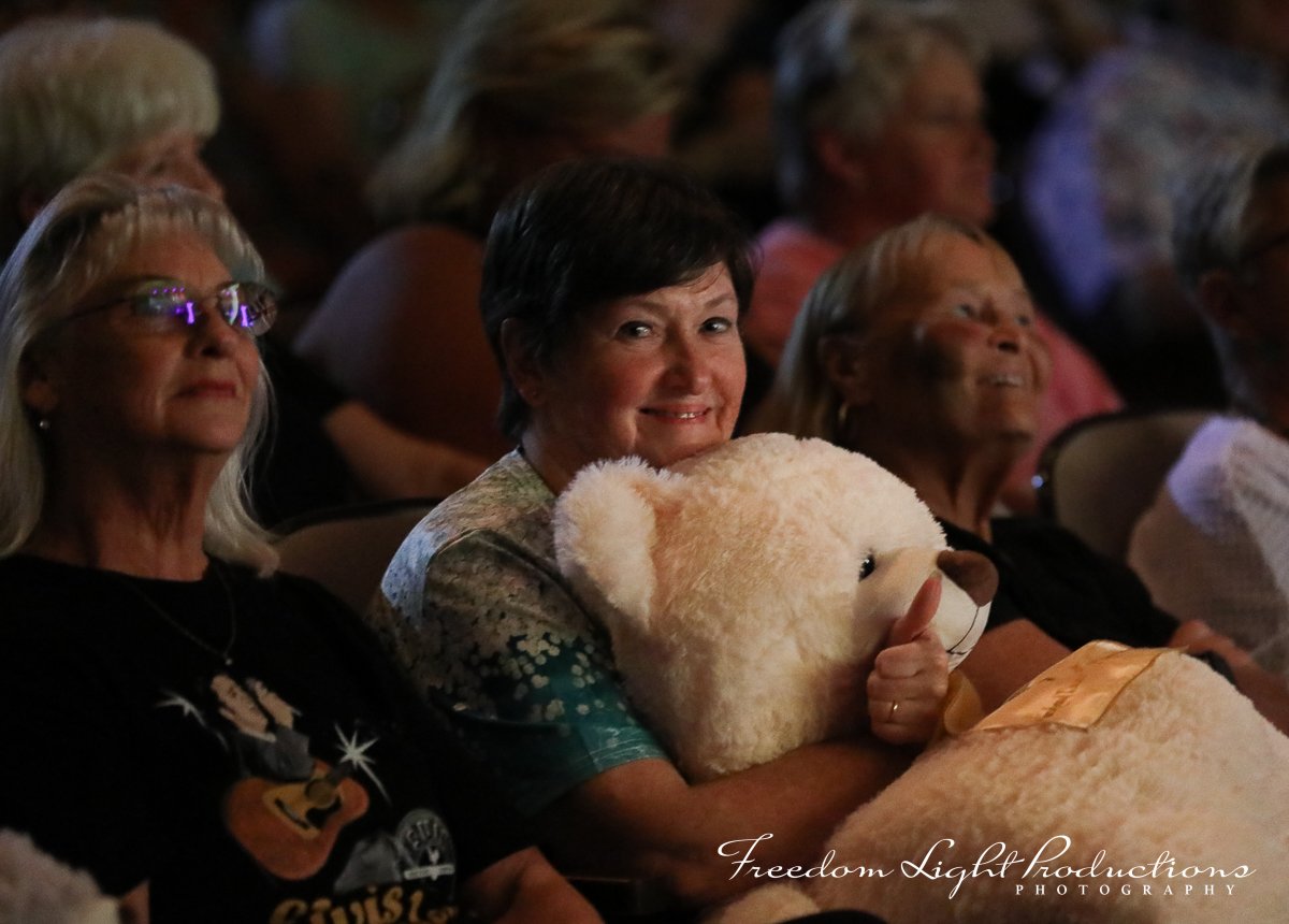 A group of women sitting in a dark room, with one woman in the center holding a large stuffed teddy bear and smiling at the camera.