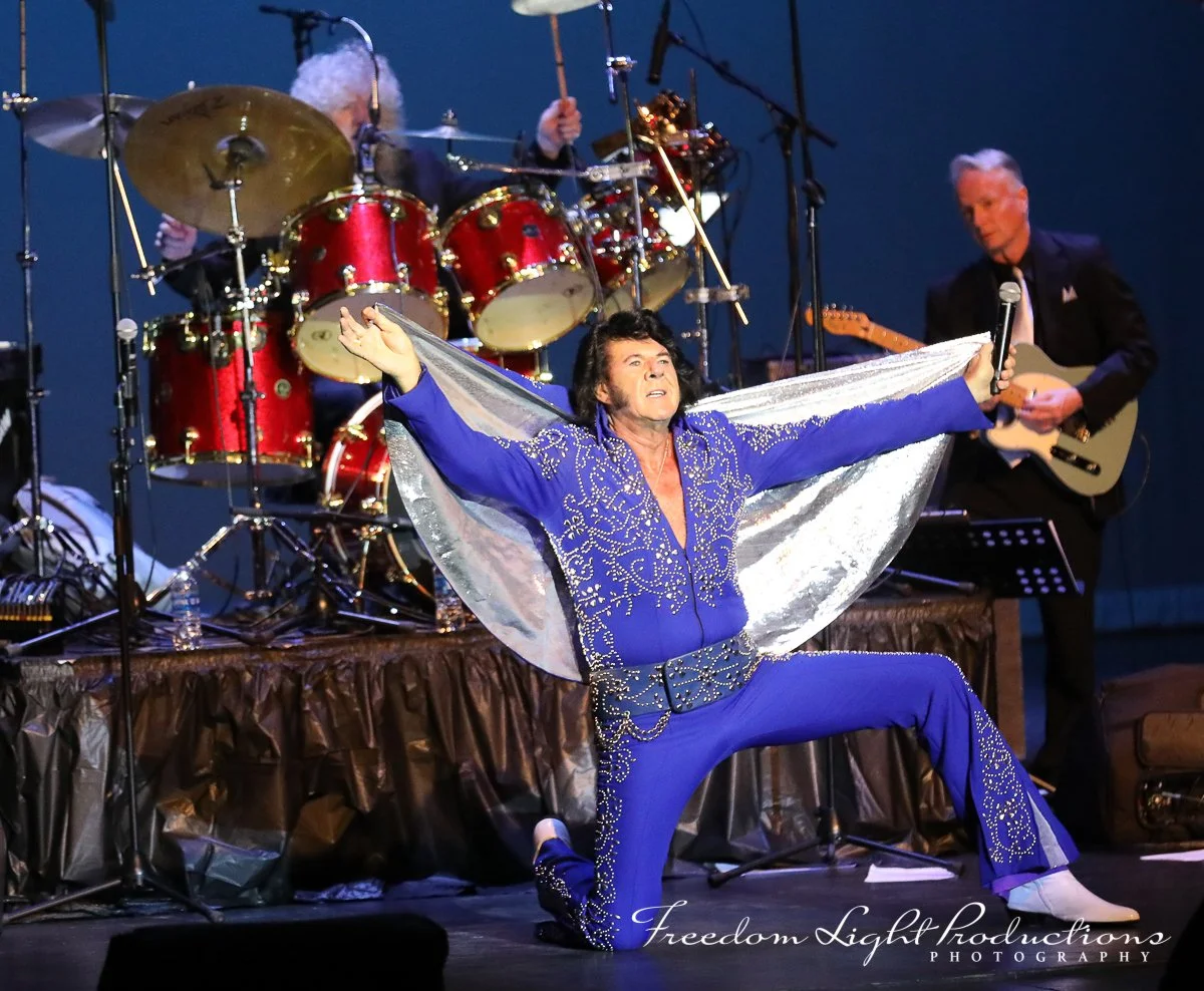 Elvis Presley impersonator performing on stage with a microphone, wearing a blue jumpsuit with rhinestone embellishments, with a band playing drums and guitar in the background.